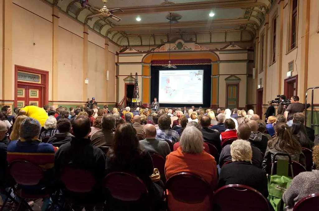 Une salle des fêtes comble pendant une réunion publique, le public face à l'écran.