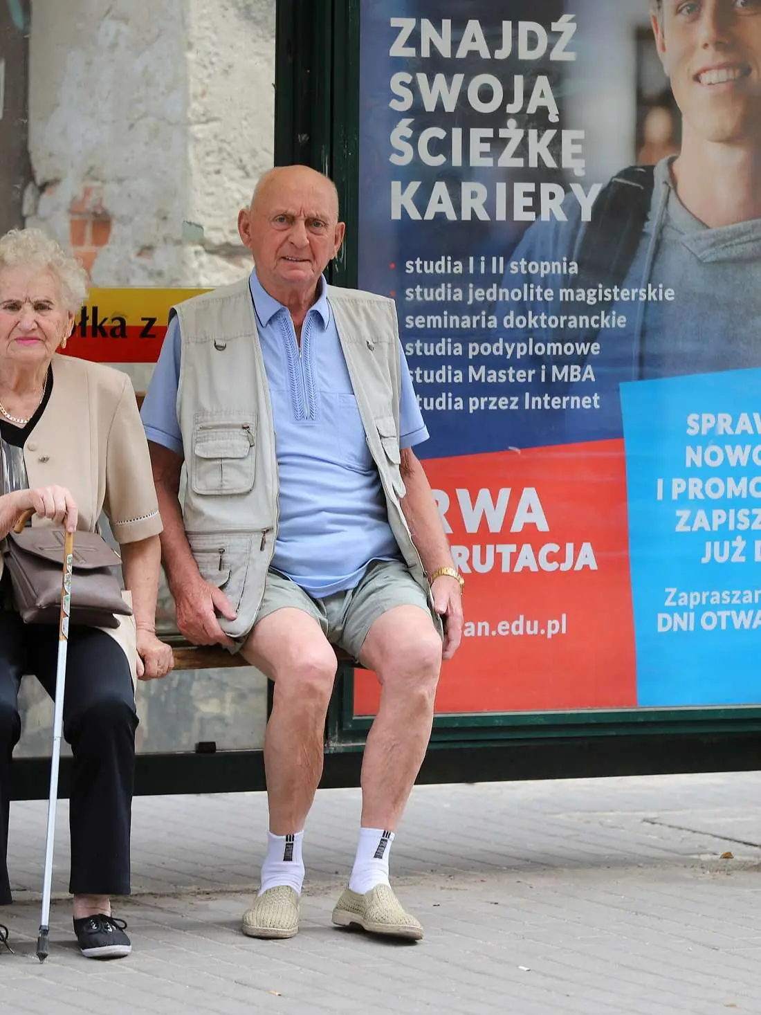 Un couple de retraités assis à un arrêt de bus, devant une grande affiche.
